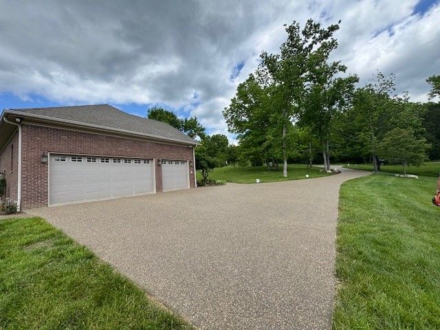 Brick garage with light gray doors and a pebble-stone driveway leading to trees under a cloudy sky.