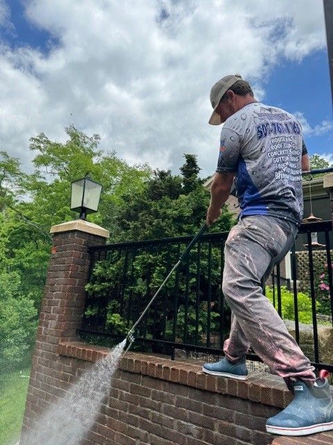 Person power washing a brick wall and black railing outdoors, cloudy sky.