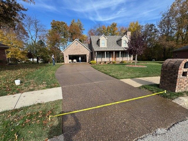 House with brown brick and gray roof. Dark driveway with yellow caution tape, lawn with trees and mailbox.
