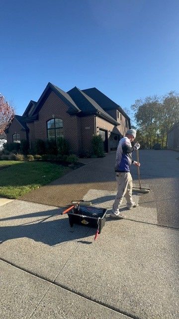 A person sweeping a driveway in front of a brick house with a dark roof on a sunny day.