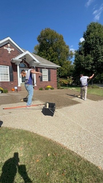 Two men working on a driveway near a brick house, one balancing on a level while others work with tools.