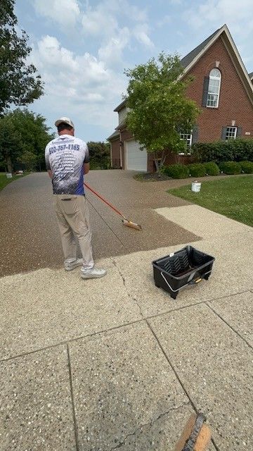Man coating a driveway with a brush. House and greenery in the background. Sunny day.