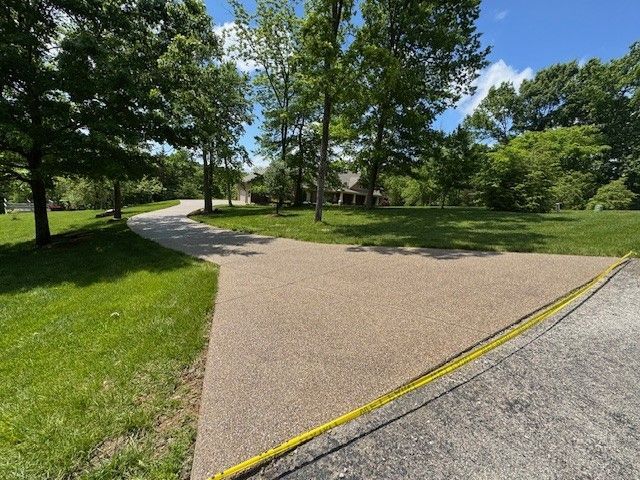 Driveway with tan surface, grassy lawn, trees, and house in the background. A yellow tape measure is visible.
