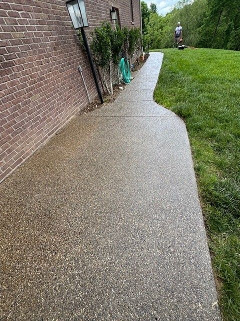 Concrete walkway next to a brick building and green lawn; a person in the distance is using a power washer.