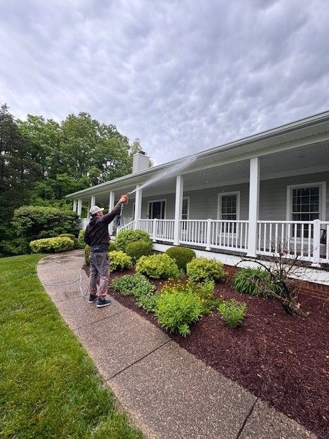 Person spraying the white siding of a house with a power washer. The house has a porch and is surrounded by greenery.