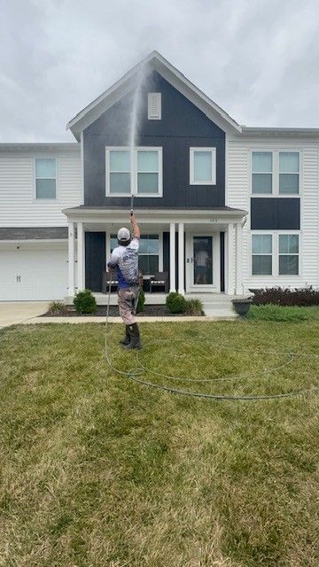 Person power washing the front of a two-story house with white siding and dark blue accents; overcast sky.