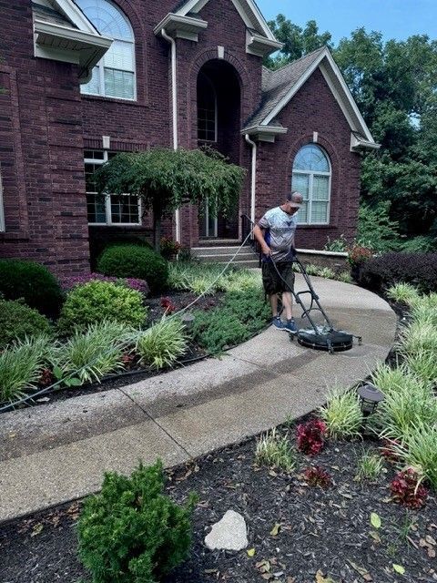 A man is cleaning the sidewalk in front of a large brick house. - Bourbon City Softwash - Louisville, KY