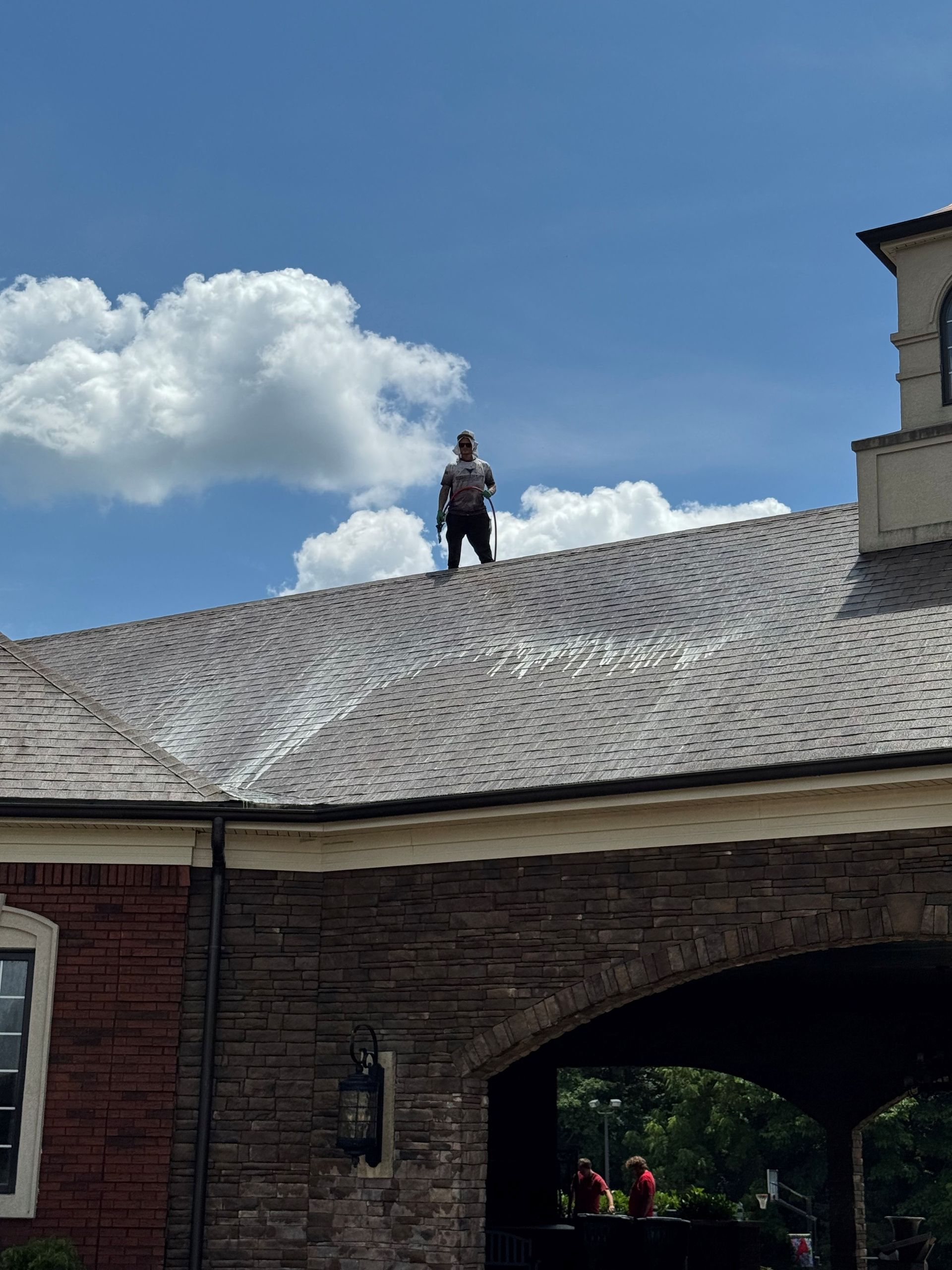 Person standing on a shingled roof, blue sky with clouds in the background. Brick building below.