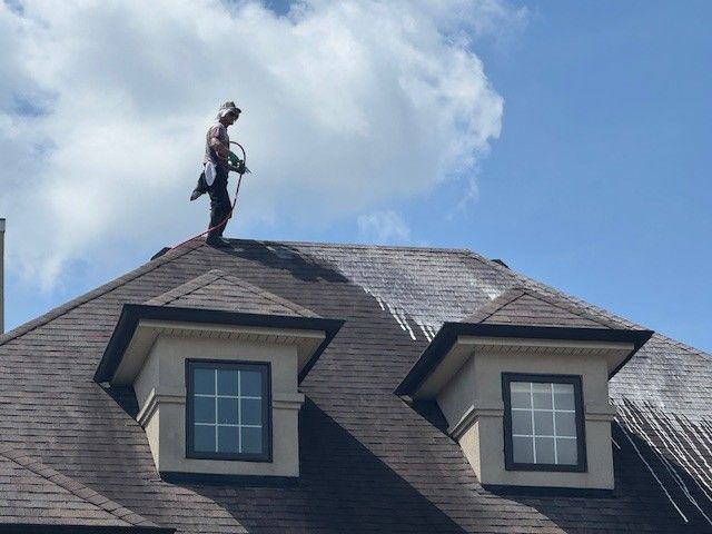 Person washing a dark brown shingled roof with a water hose under a cloudy sky.