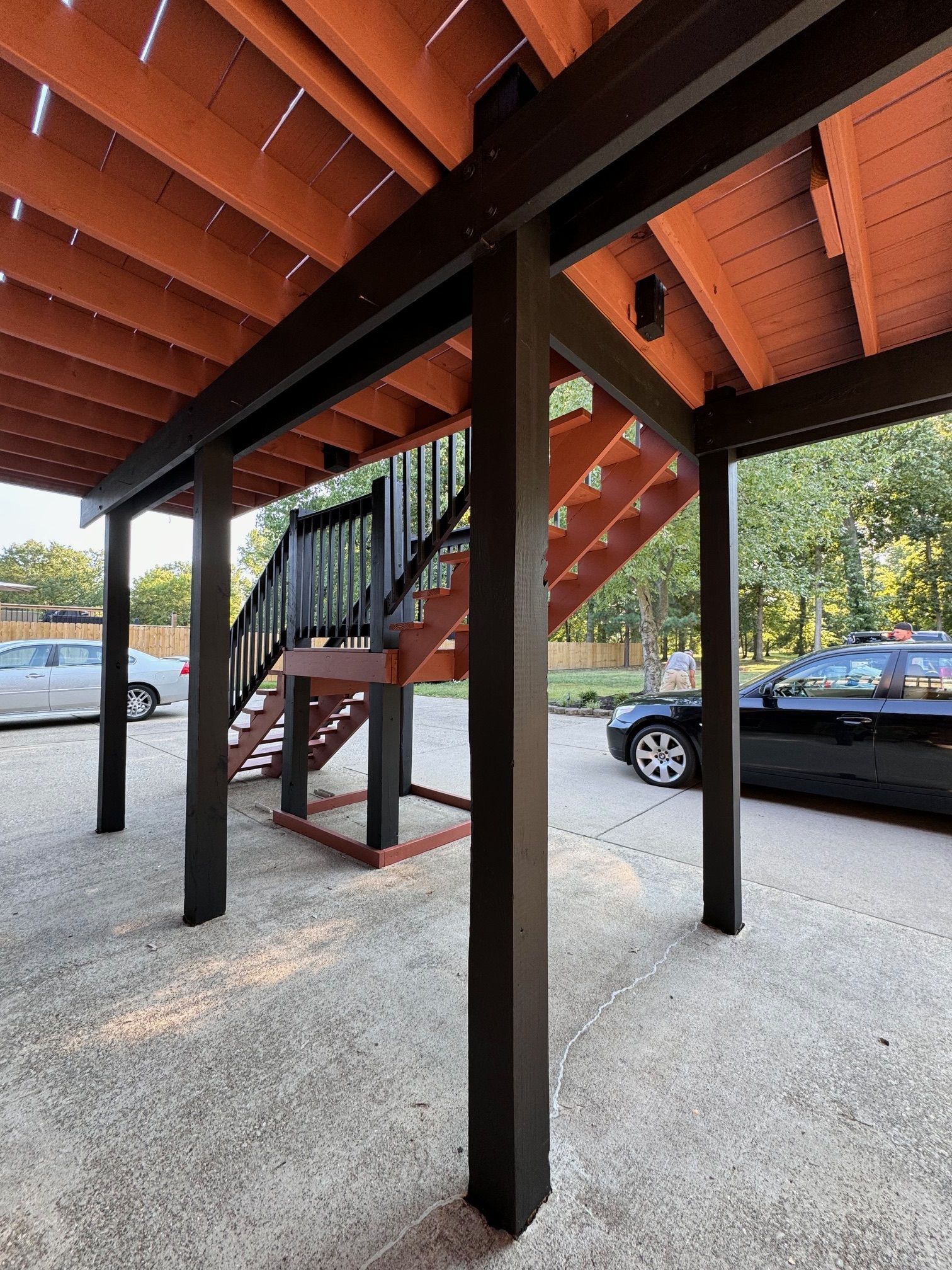 A car is parked under a wooden deck with stairs leading up to it.