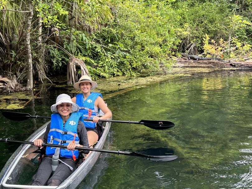 Two people kayaking on clear water, wearing life vests and hats, near lush green vegetation.