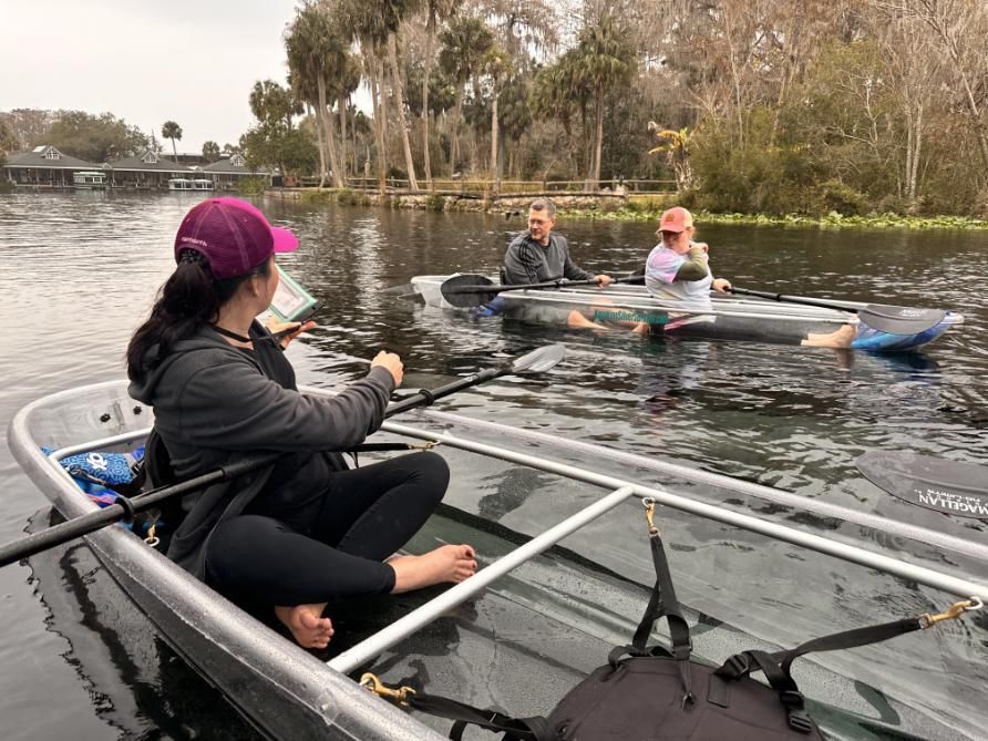 Woman in clear kayak paddles; two others relax in kayaks on a calm river; trees and buildings in the background.