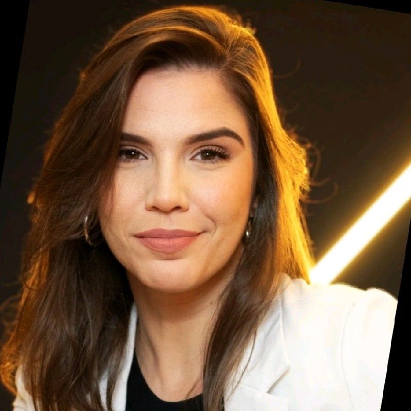Mulher de cabelos castanhos, vestindo um blazer branco, sorrindo para a câmera. Contraluz, com luz amarela.