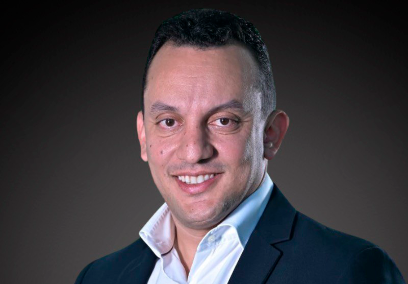 Homem de terno escuro e camisa branca sorri, retrato contra um fundo marrom.