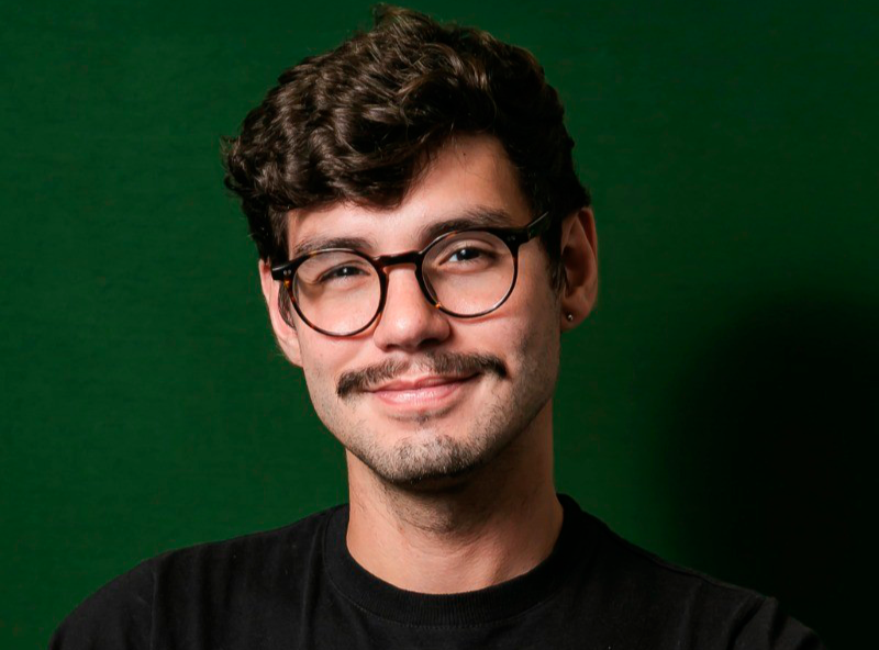 Homem de cabelos escuros e encaracolados, bigode e óculos, sorrindo em frente a um fundo verde.