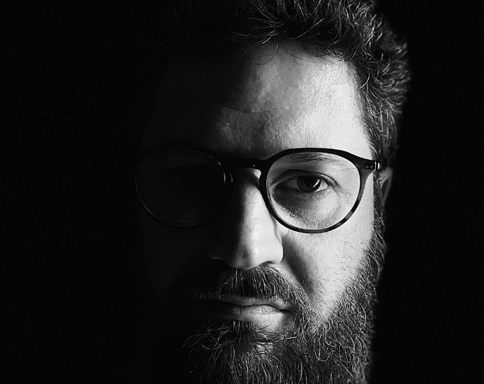 Man with glasses and beard, partially lit, looking at the camera.