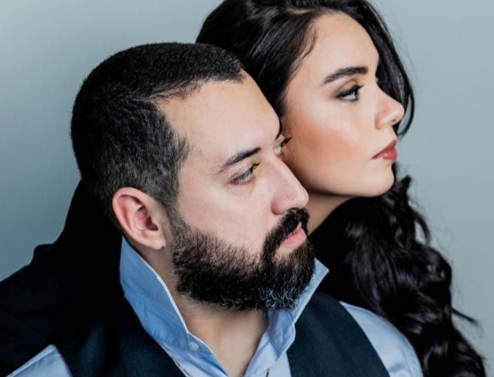 Homem com barba e mulher com cabelo escuro posando juntos; ambos olhando para longe.