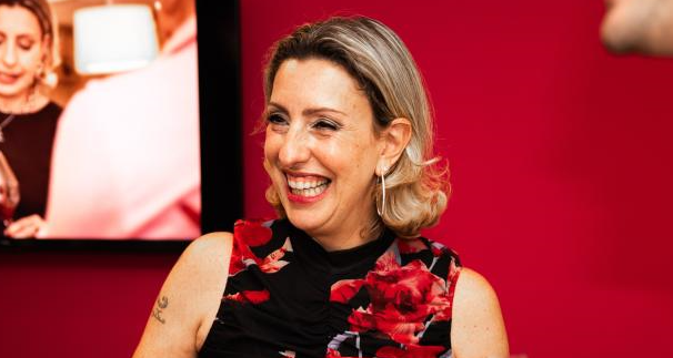 A person with blonde hair in a floral top smiles warmly in front of a red wall.