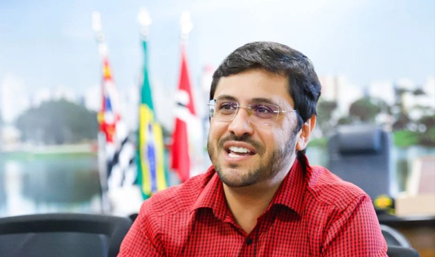 Homem de camisa vermelha e óculos, sorrindo, falando em um escritório com bandeiras ao fundo.