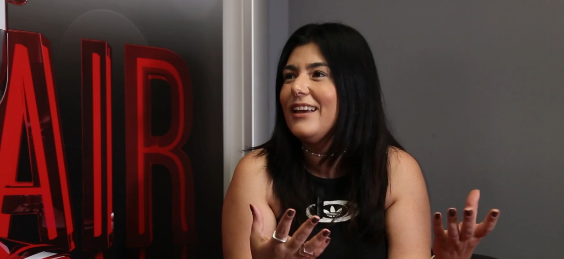 Mulher falando e gesticulando com as mãos; sentada em frente a um painel com a palavra 