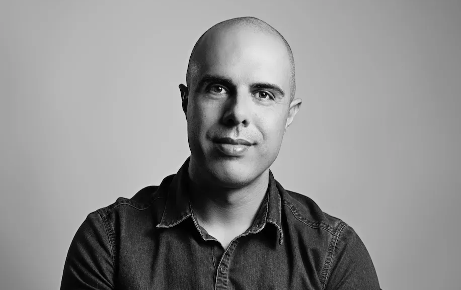 A black-and-white portrait of a person with a shaved head, wearing a button-up shirt, looking directly at the camera.