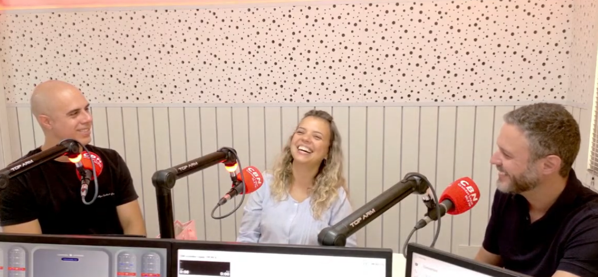 Três pessoas sentam-se à mesa de um estúdio com microfones, rindo durante a gravação de um podcast em uma sala de cores claras.