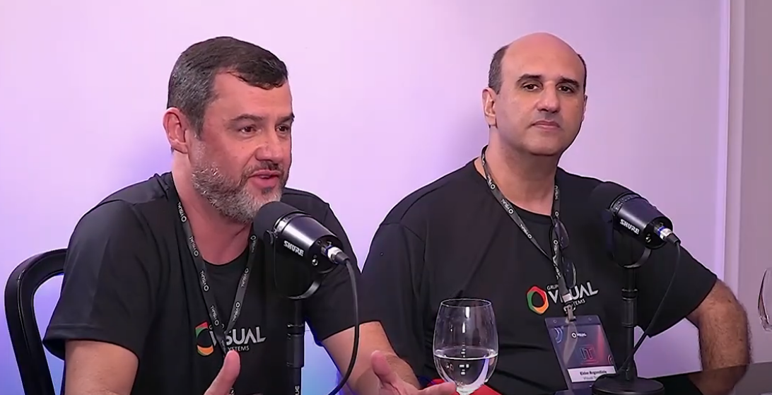 Dois homens de camisa preta com microfones, sentados. Um fala, o outro observa. Em um ambiente interno.