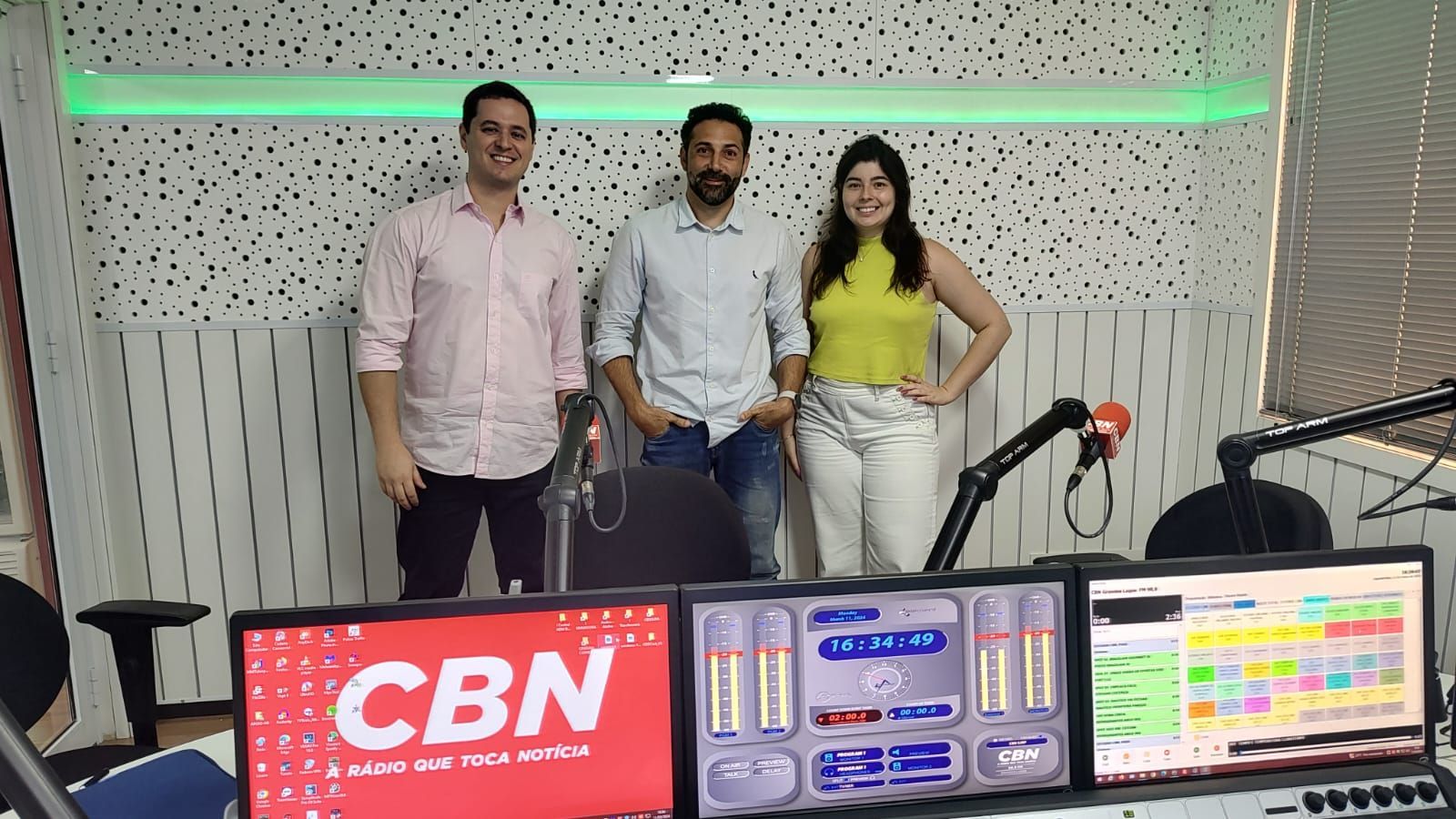 Três pessoas estão em frente a uma placa da CBN em um estúdio de rádio.
