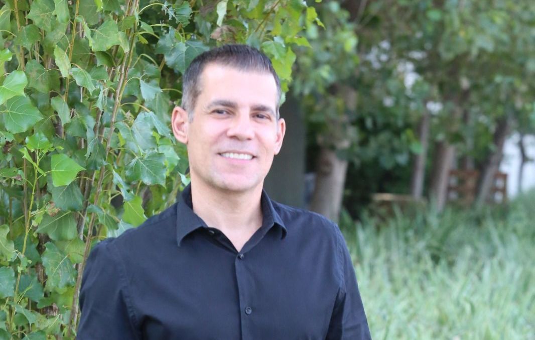 Um homem de camisa preta e calça jeans está parado na frente de um arbusto.