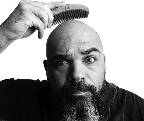 Homem penteia a cabeça calva, de olhos arregalados, com um pente de madeira. Preto e branco.