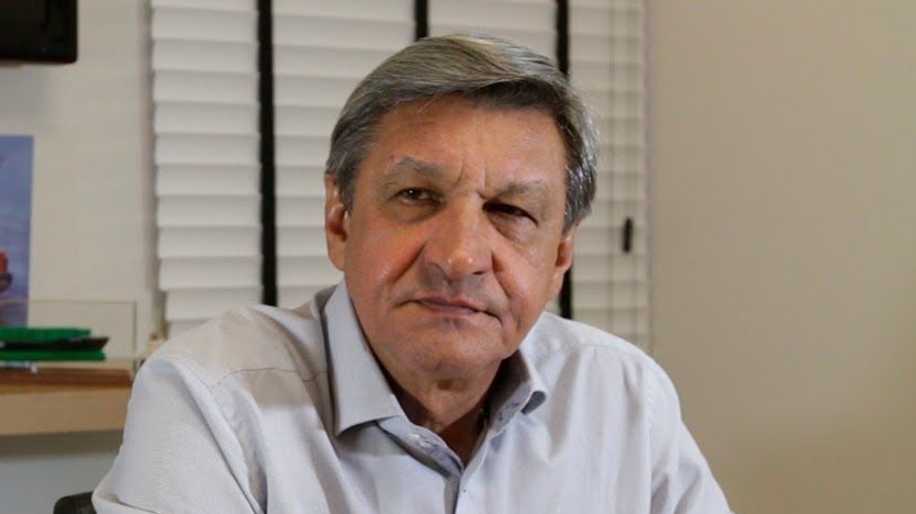 Homem de cabelos grisalhos e camisa olhando para a câmera, em ambiente interno, próximo a persianas.