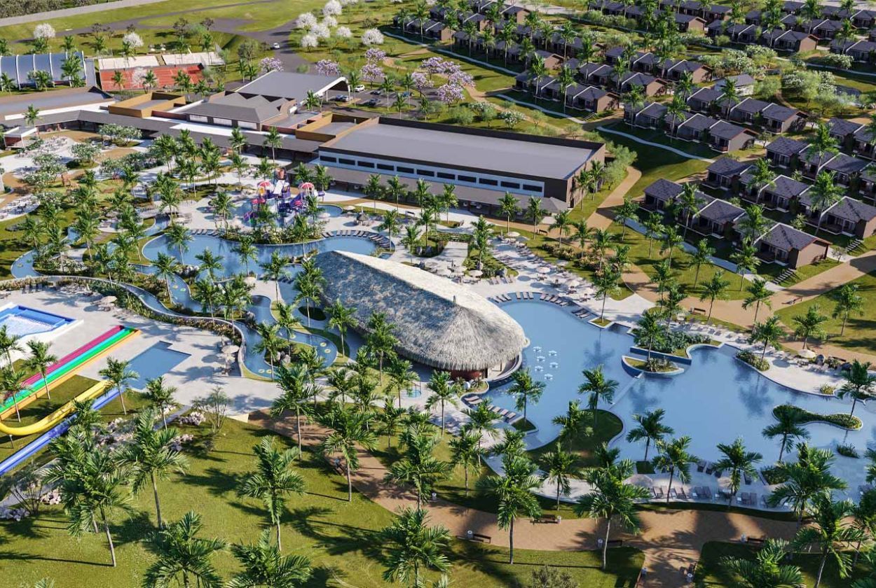 Aerial view of a resort with a large pool, cabana, and buildings with green surroundings and palm trees.