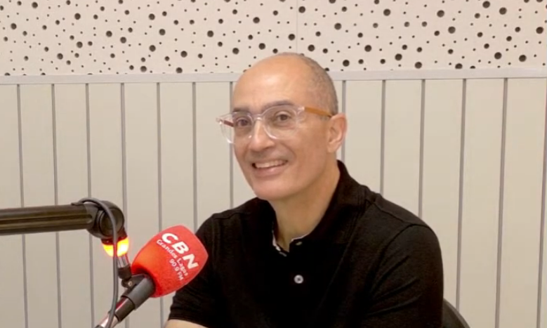 A smiling person in a black polo shirt speaks into a red CBN microphone in a studio with light-colored paneled walls.