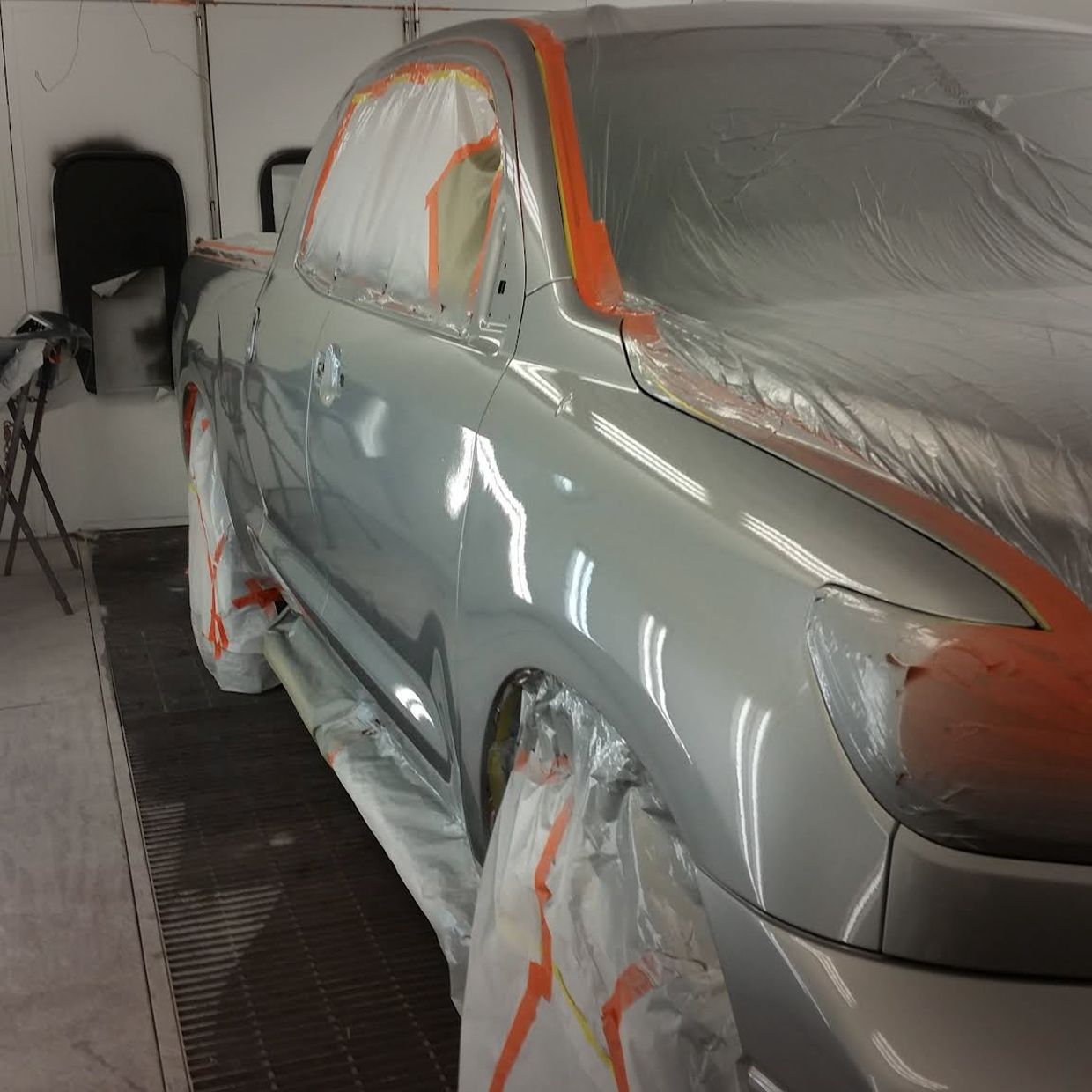Silver car in a paint booth, partially covered with plastic and orange tape, ready for painting. | Stu's Collision Center