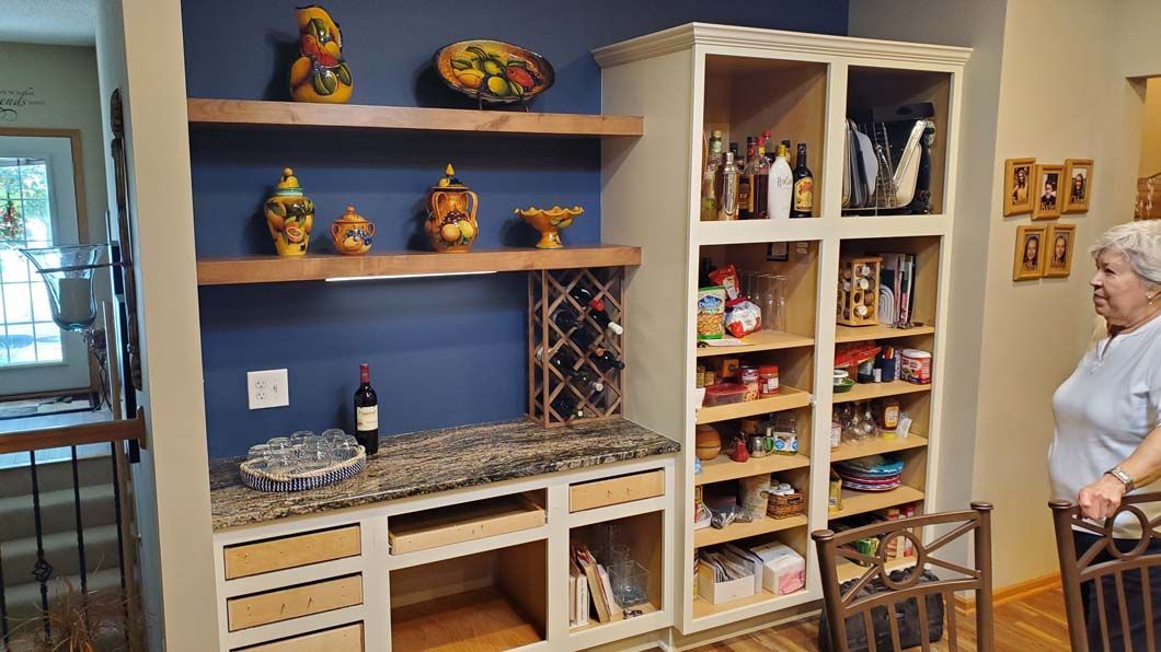 A woman is standing in front of a kitchen with shelves and a wine rack.