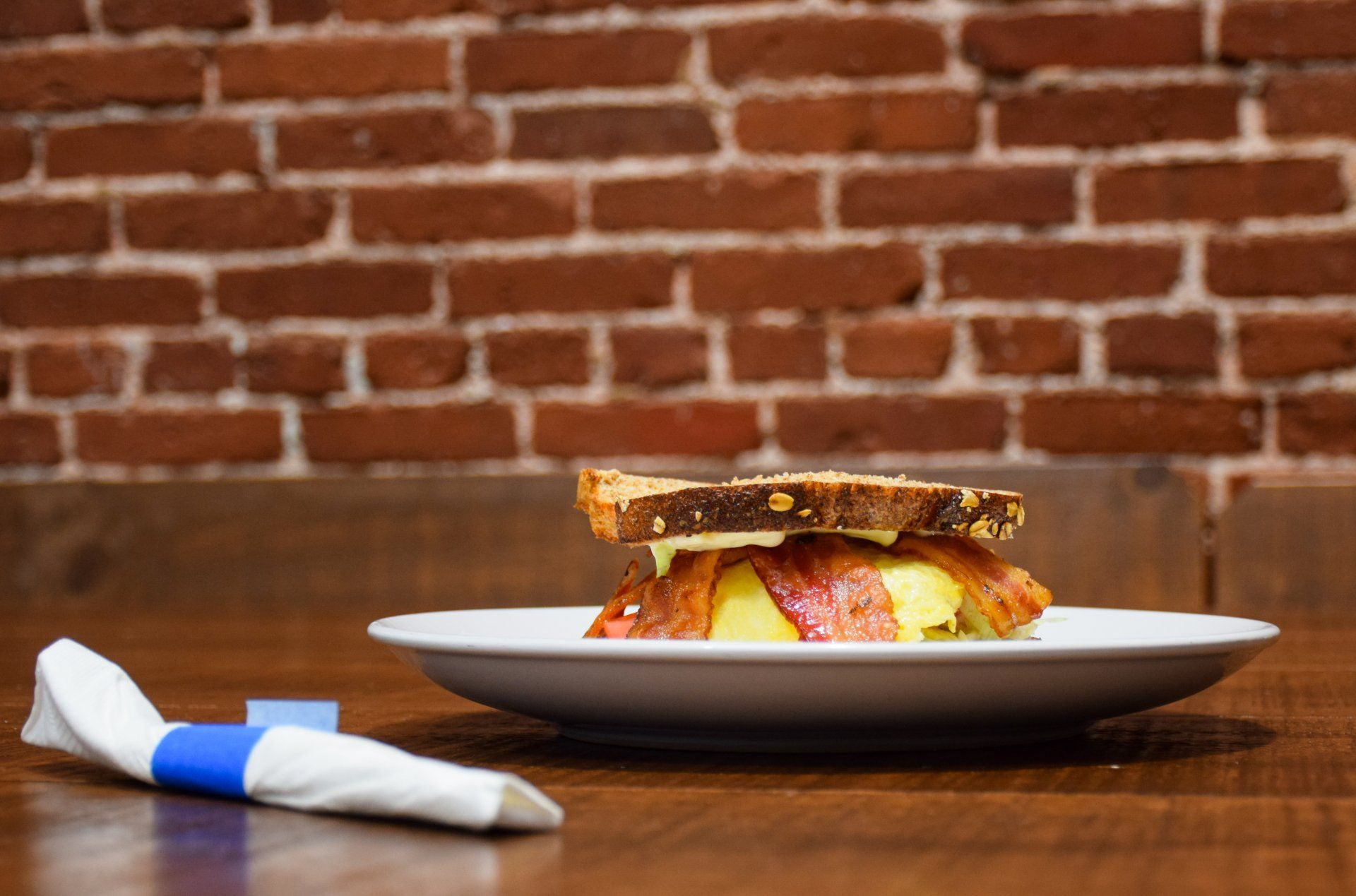 A sandwich on a plate with a napkin and a brick wall in the background.