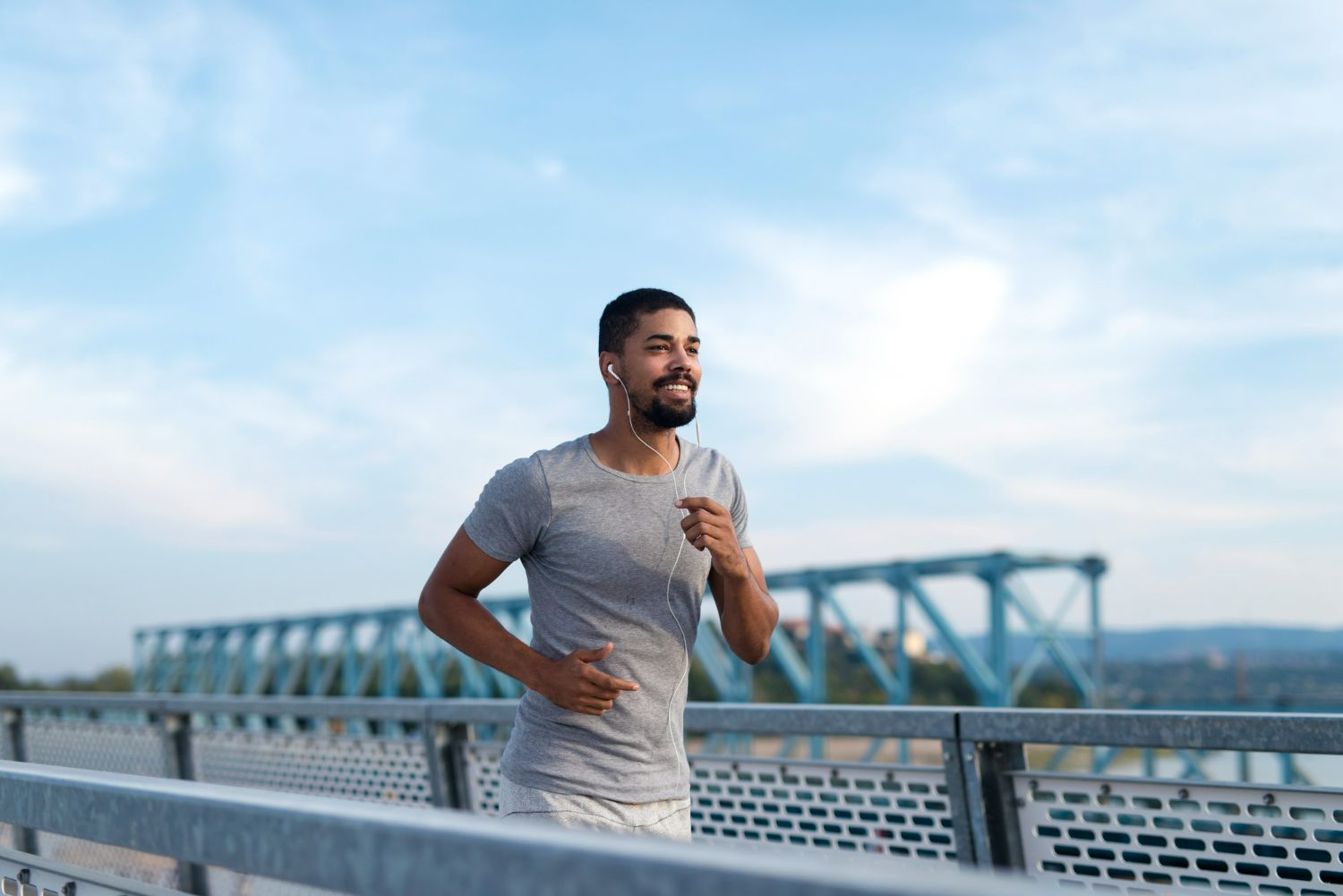 Man running on a bridge, smiling, wearing headphones, and gray athletic wear. Blue sky in the background.