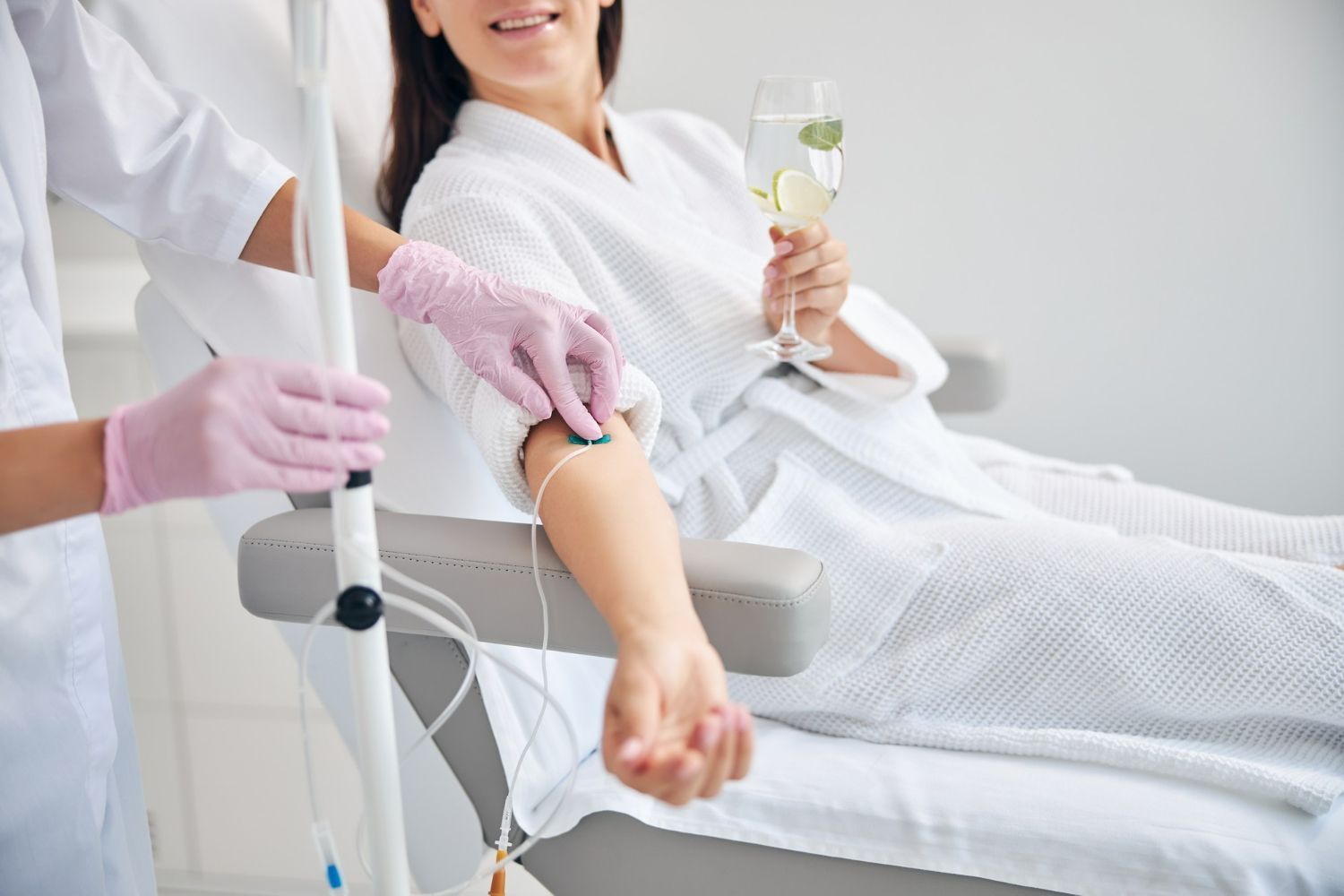 Woman receiving IV drip, wearing robe, holding champagne glass, smiling.
