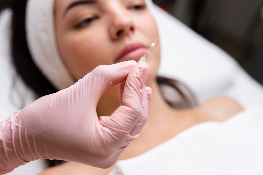 Close-up of a gloved hand holding a syringe near a woman's lips, pre-injection, white setting.