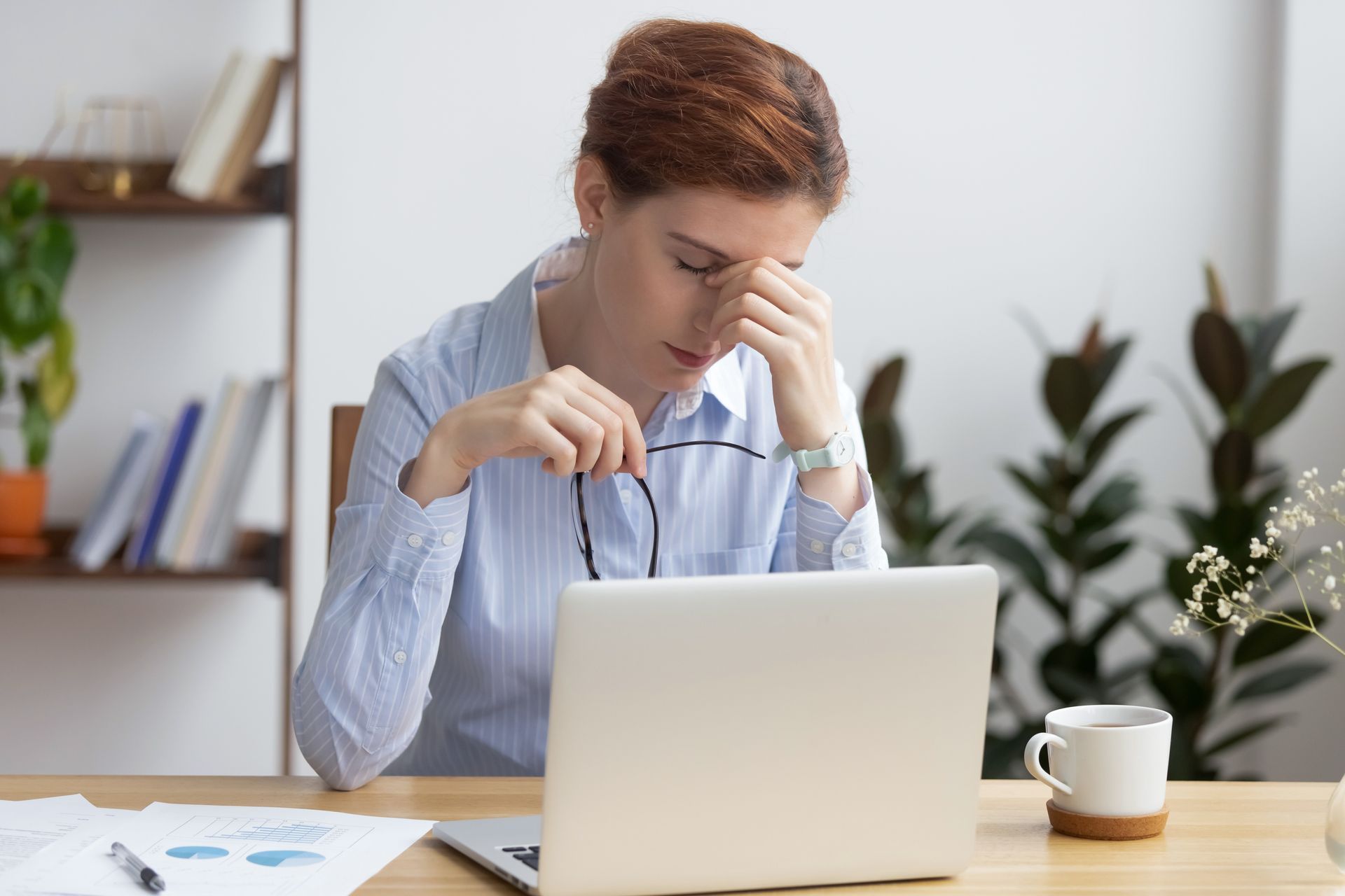 Woman in office, frustrated, holding glasses, touching her eyes in front of a laptop.