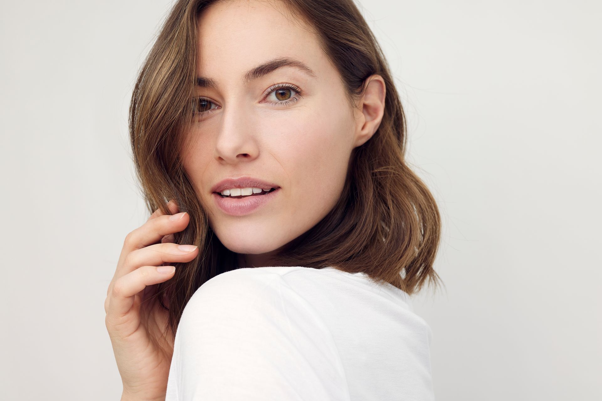 Woman with brown hair, smiling, hand near face, white shirt, neutral background.
