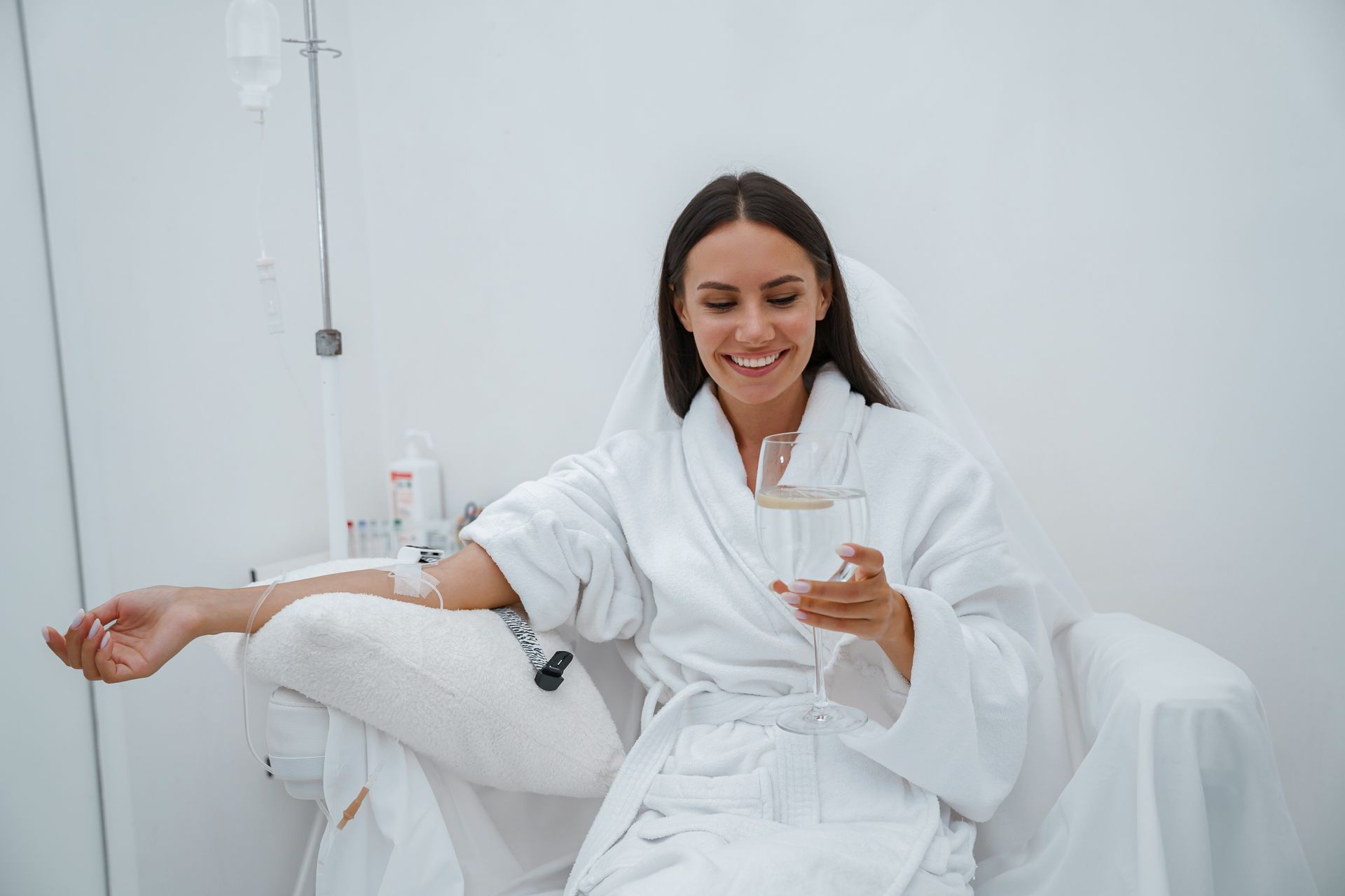 Woman in white robe getting IV drip, holding a glass, smiling in a bright room.