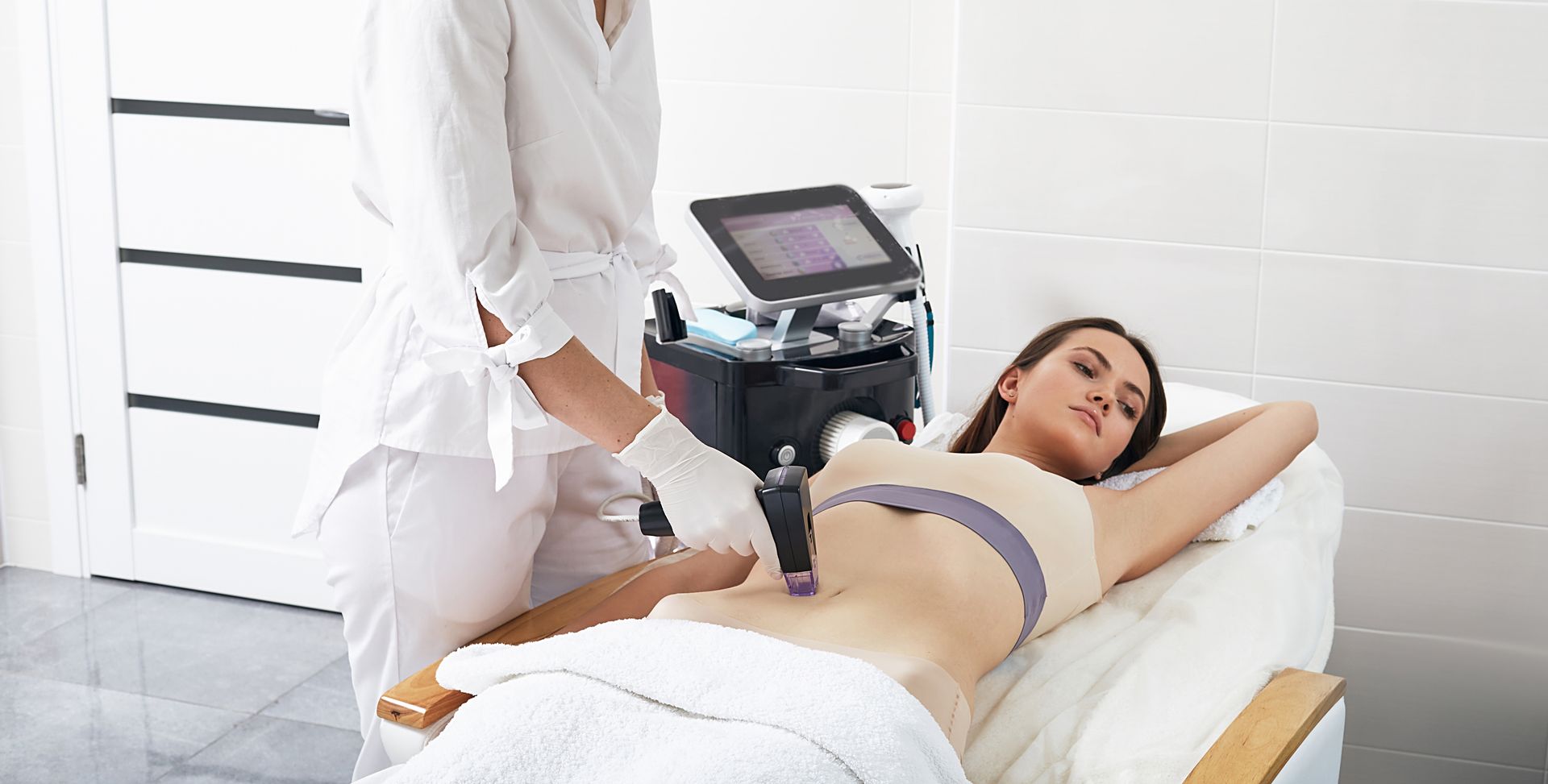 Woman receiving body contouring treatment on her abdomen in a clinic. A technician uses a device, lying on a white bed.