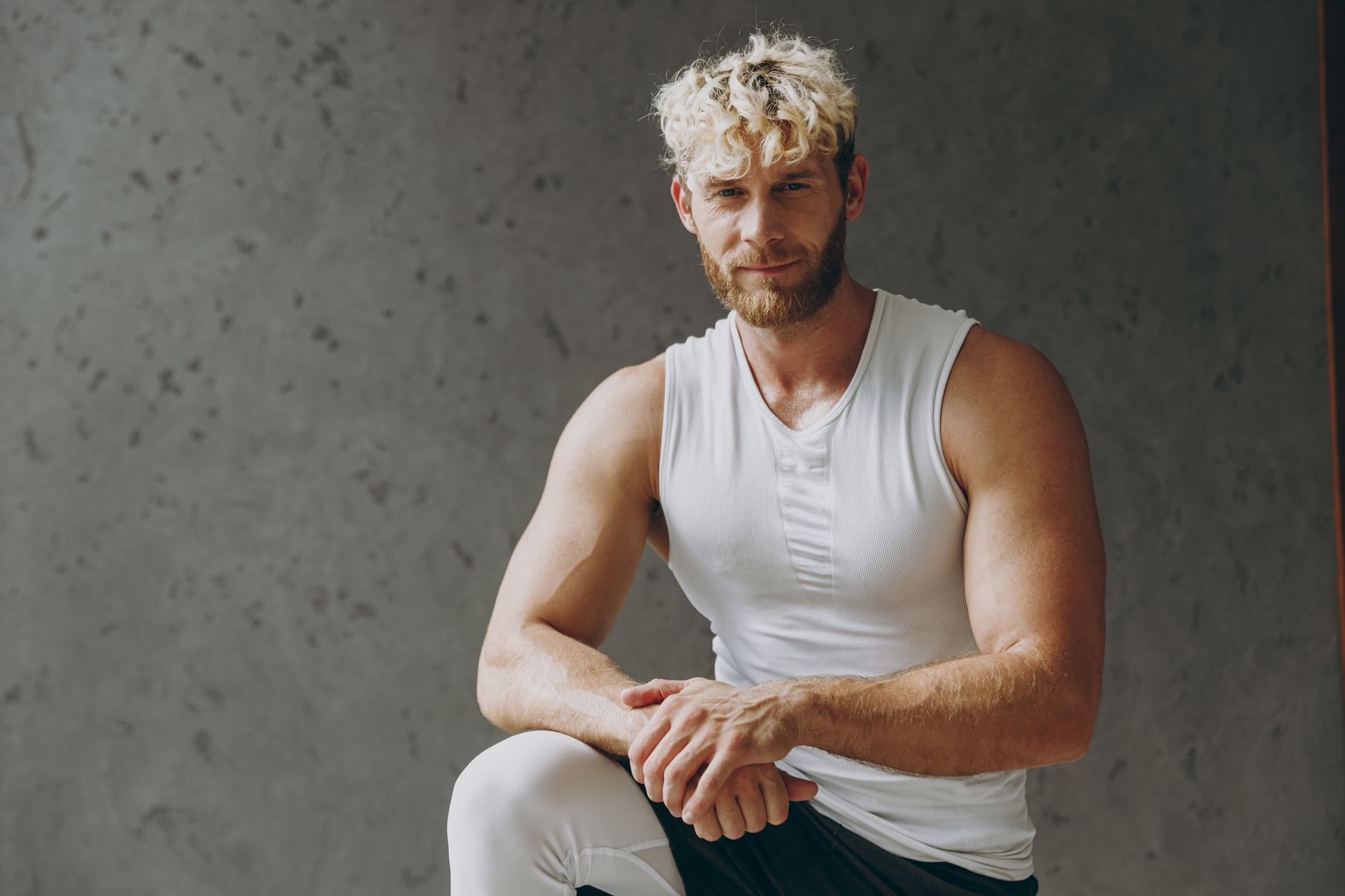 Man with blonde hair and beard in white tank top and leggings, sitting, looking at camera.