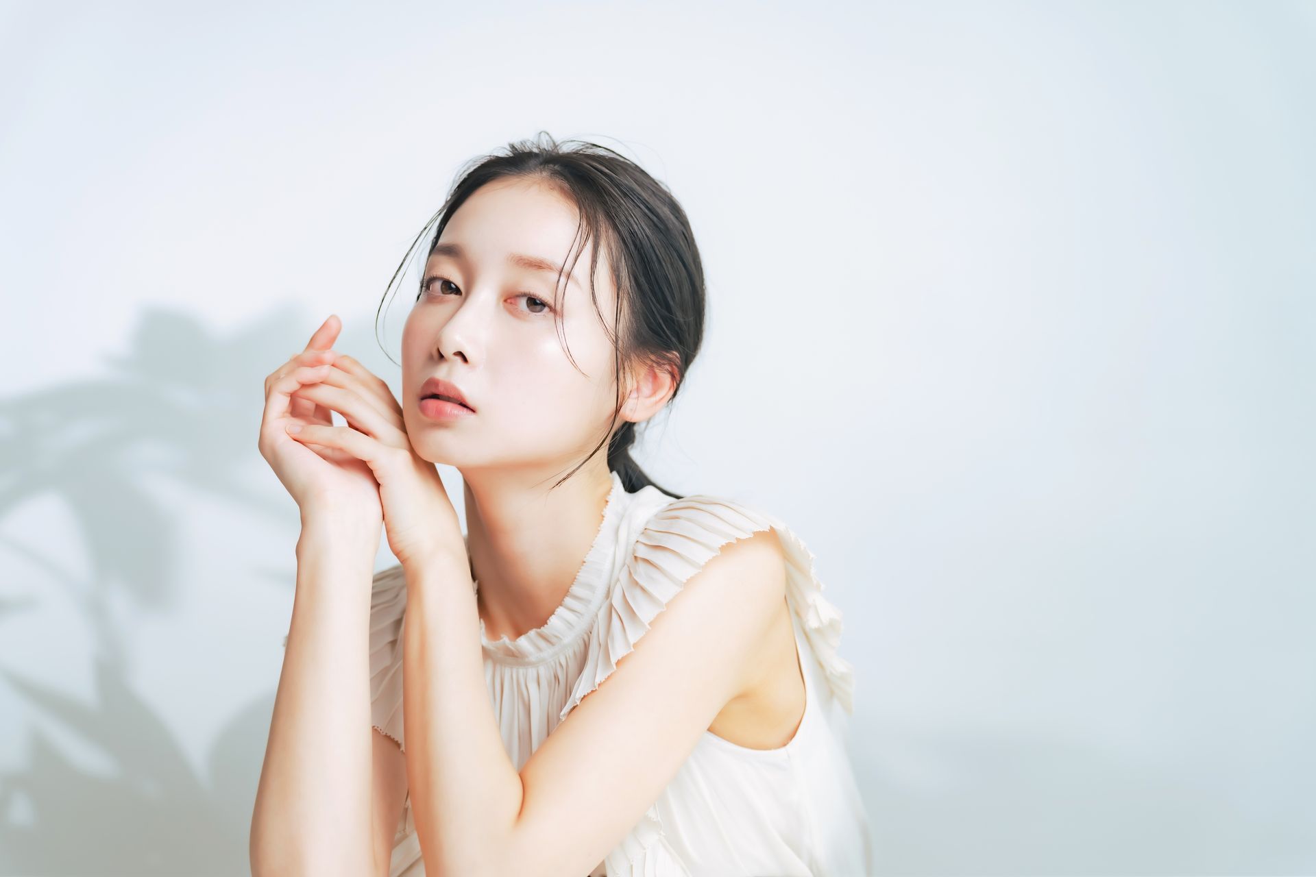 Woman with dark hair, pale skin, wearing a white blouse, poses in a studio with a shadow of a plant on the wall.