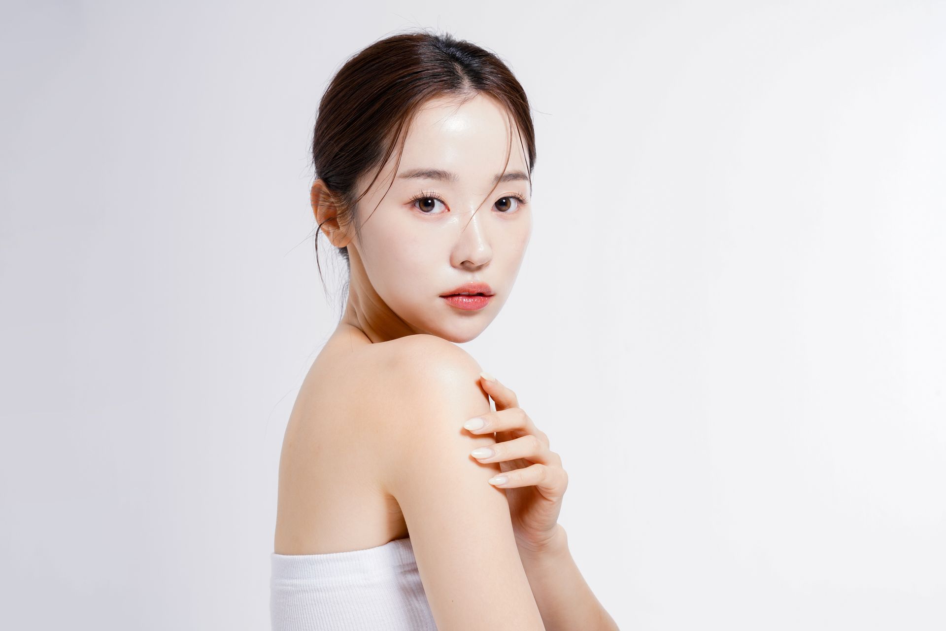 Woman looking over her shoulder, touching her shoulder, soft lighting, white background.