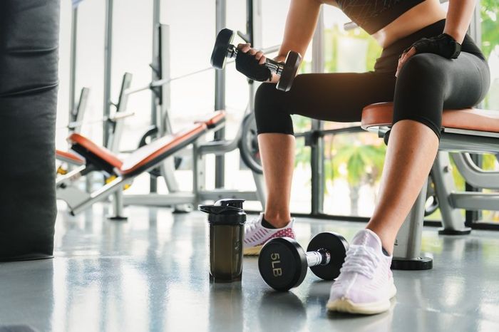 A woman is sitting on a bench in a gym holding a dumbbell.