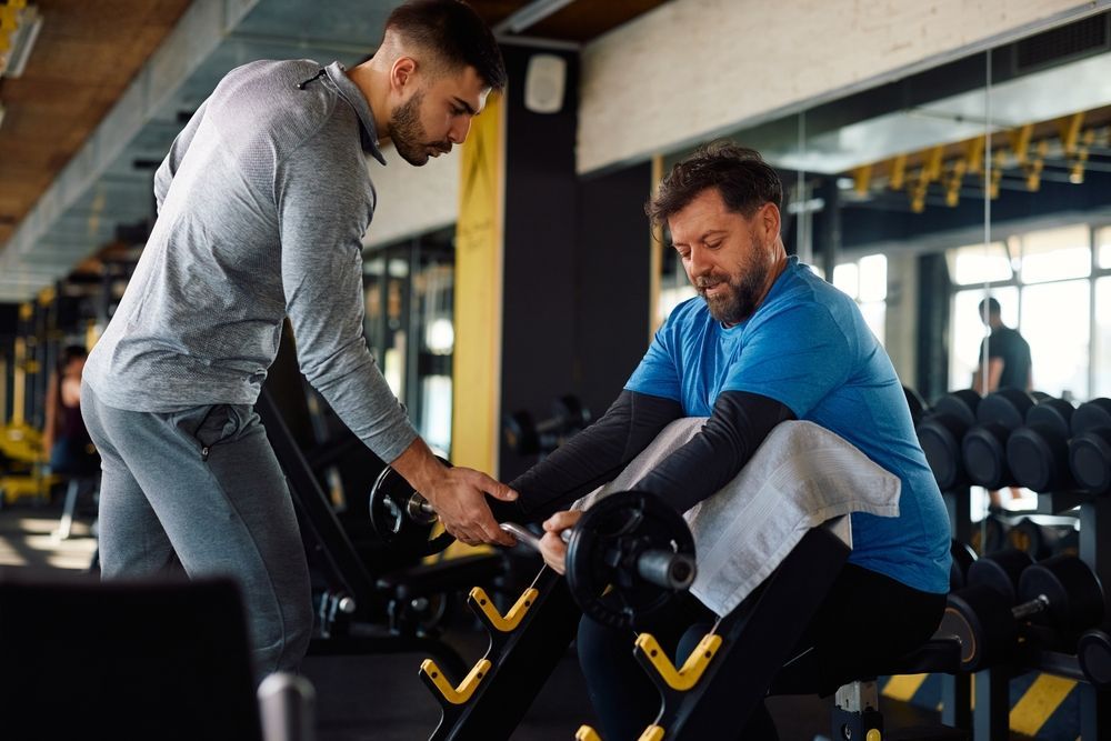 A coach is helping another man lift weights in a gym.