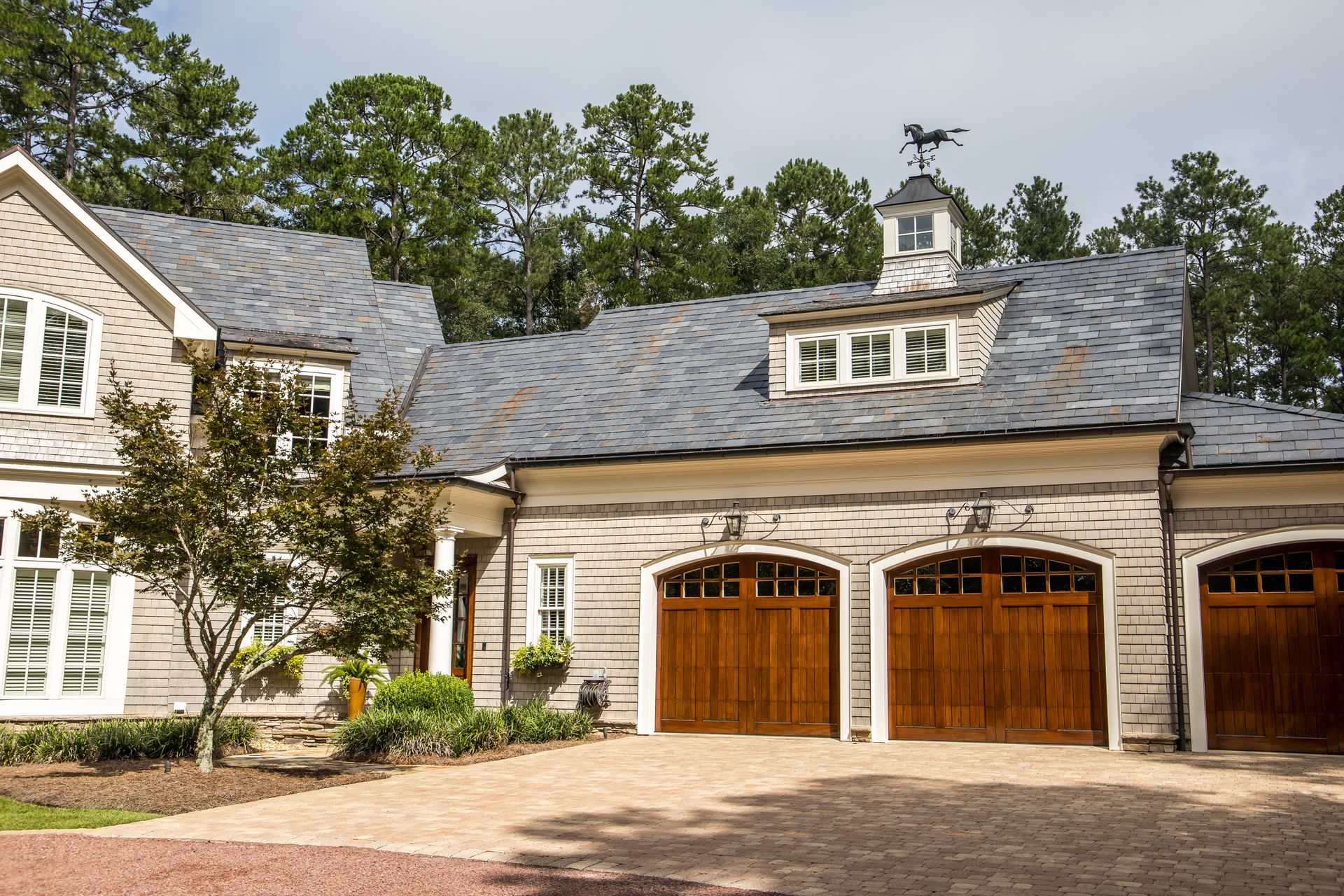 Stone house exterior with gray roof, arched wooden double garage doors, and a paved driveway surrounded by trees