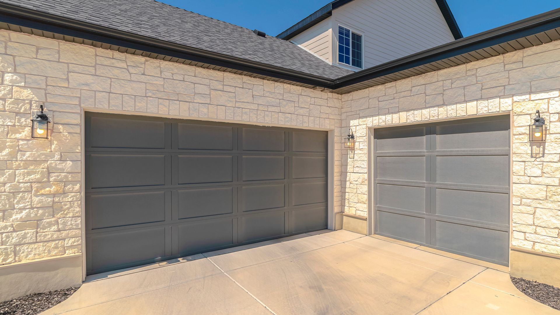 Exterior of a house with two gray garage doors and a stone facade in sunlight